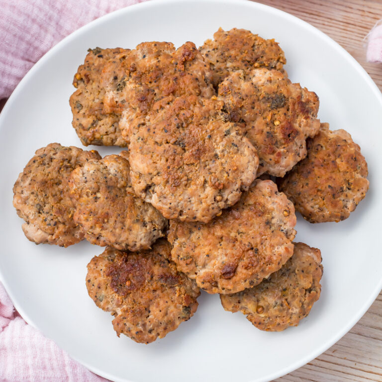 A white plate filled with several cooked, seasoned turkey breakfast sausage patties sits on a light wooden surface.