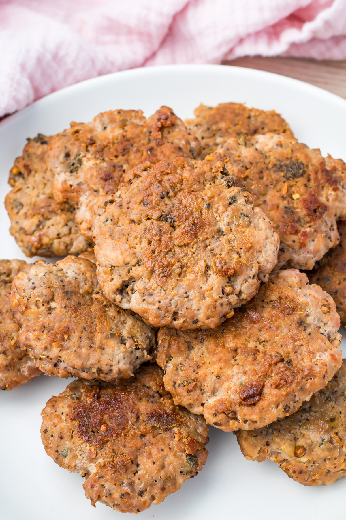 A white plate holds several browned, homemade turkey breakfast sausage patties, stacked closely together. A pink cloth is partially visible in the background.