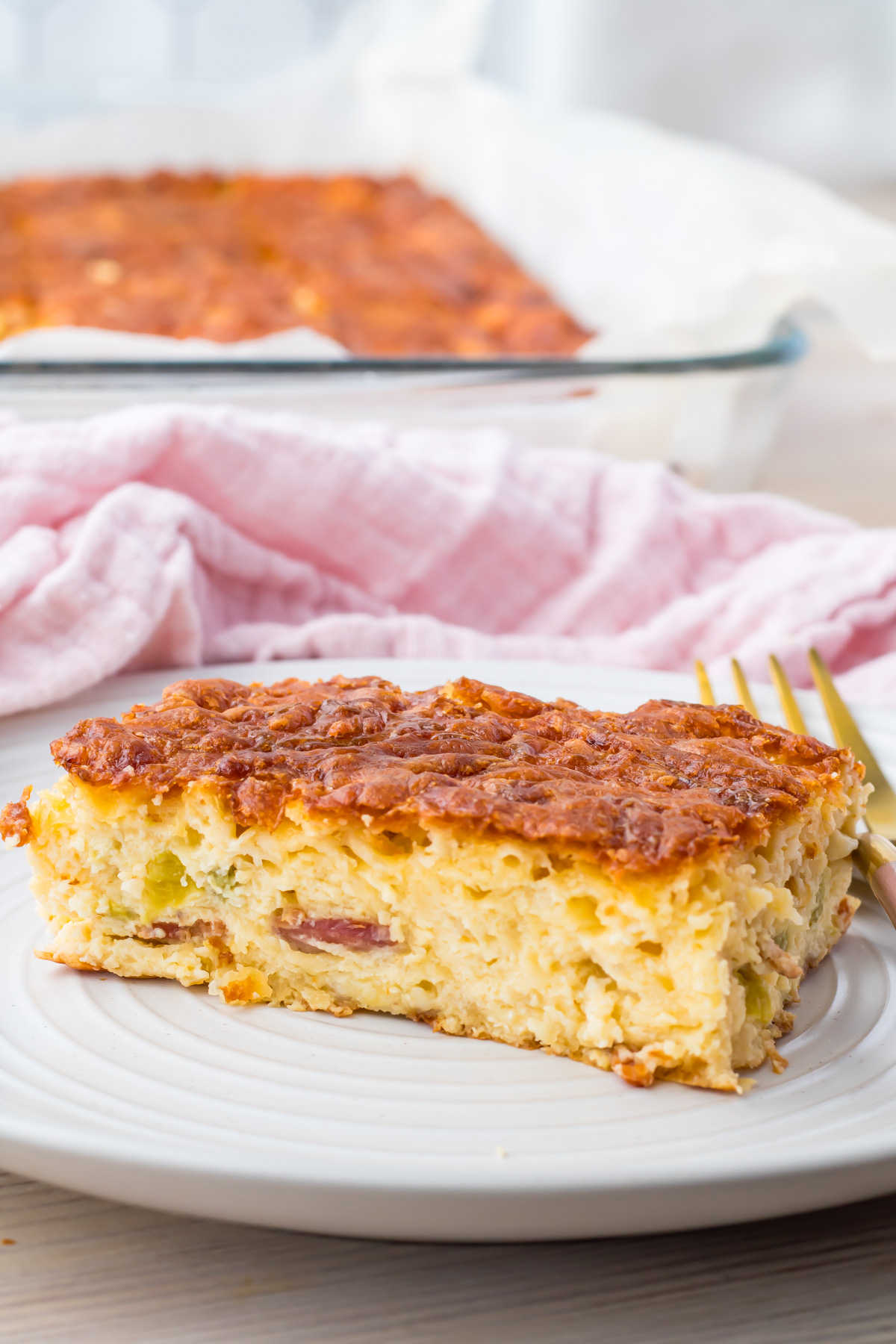 A slice of baked cottage cheese egg casserole with visible pieces of meat and vegetables sits on a plate, with the remaining casserole in a glass dish in the background.