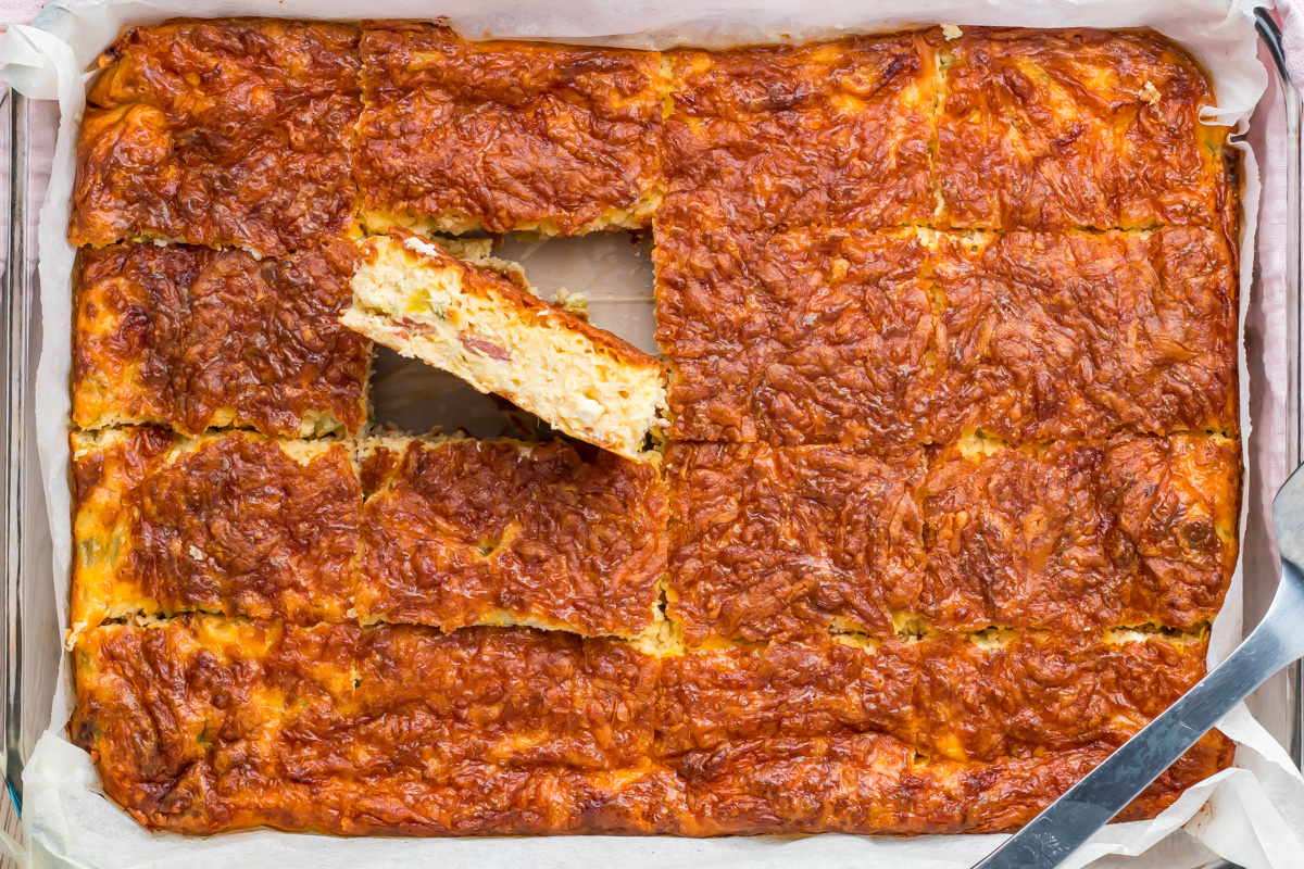 A baked cottage cheese egg casserole with a golden, cheesy crust is sliced into squares, with one piece partially removed from the pan and resting on its side.