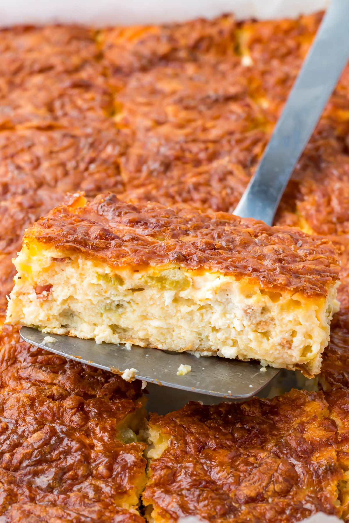 A metal spatula lifts a square slice of cottage cheese egg casserole with a browned, cheesy top from a pan of similar pieces.