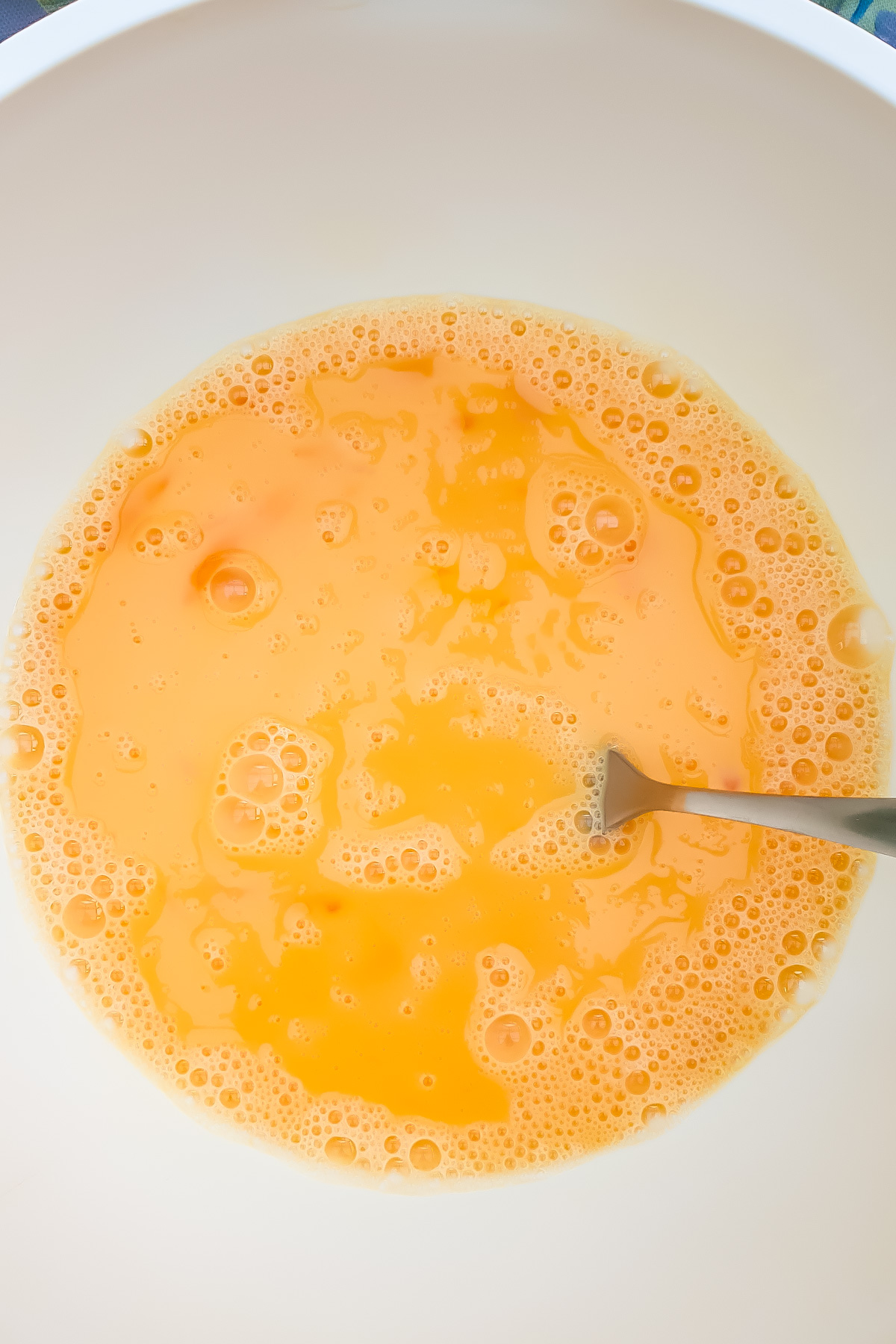 A metal fork in a white bowl with beaten eggs, showing a yellow-orange liquid with bubbles on the surface.