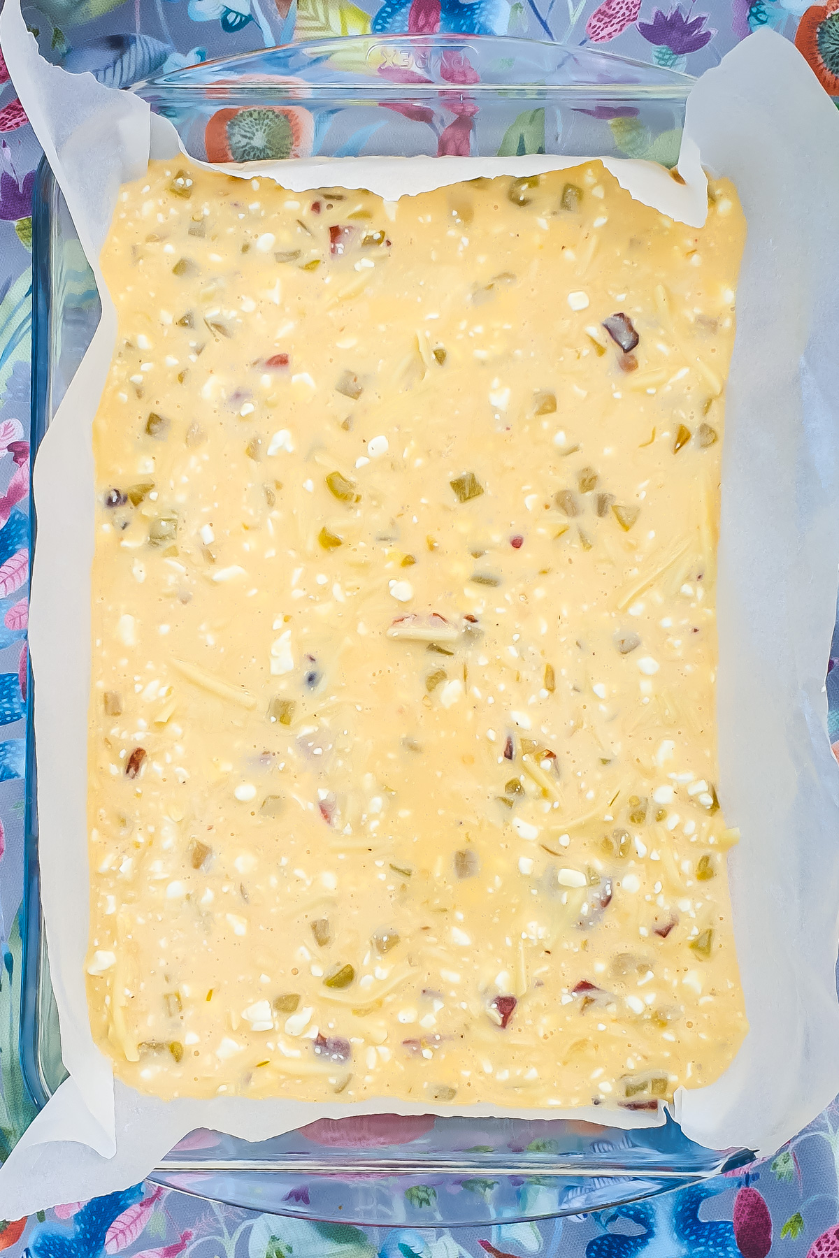 A rectangular baking dish lined with parchment paper, filled with an uncooked egg casserole batter featuring visible chunks of vegetables and cottage cheese.