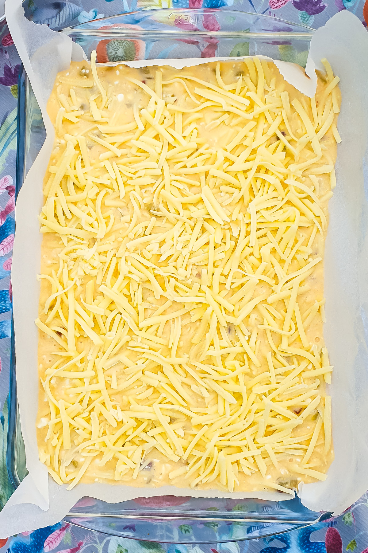 A glass baking dish lined with parchment paper holds an uncooked Cottage Cheese Casserole batter topped with shredded cheese, set on a floral-patterned surface.