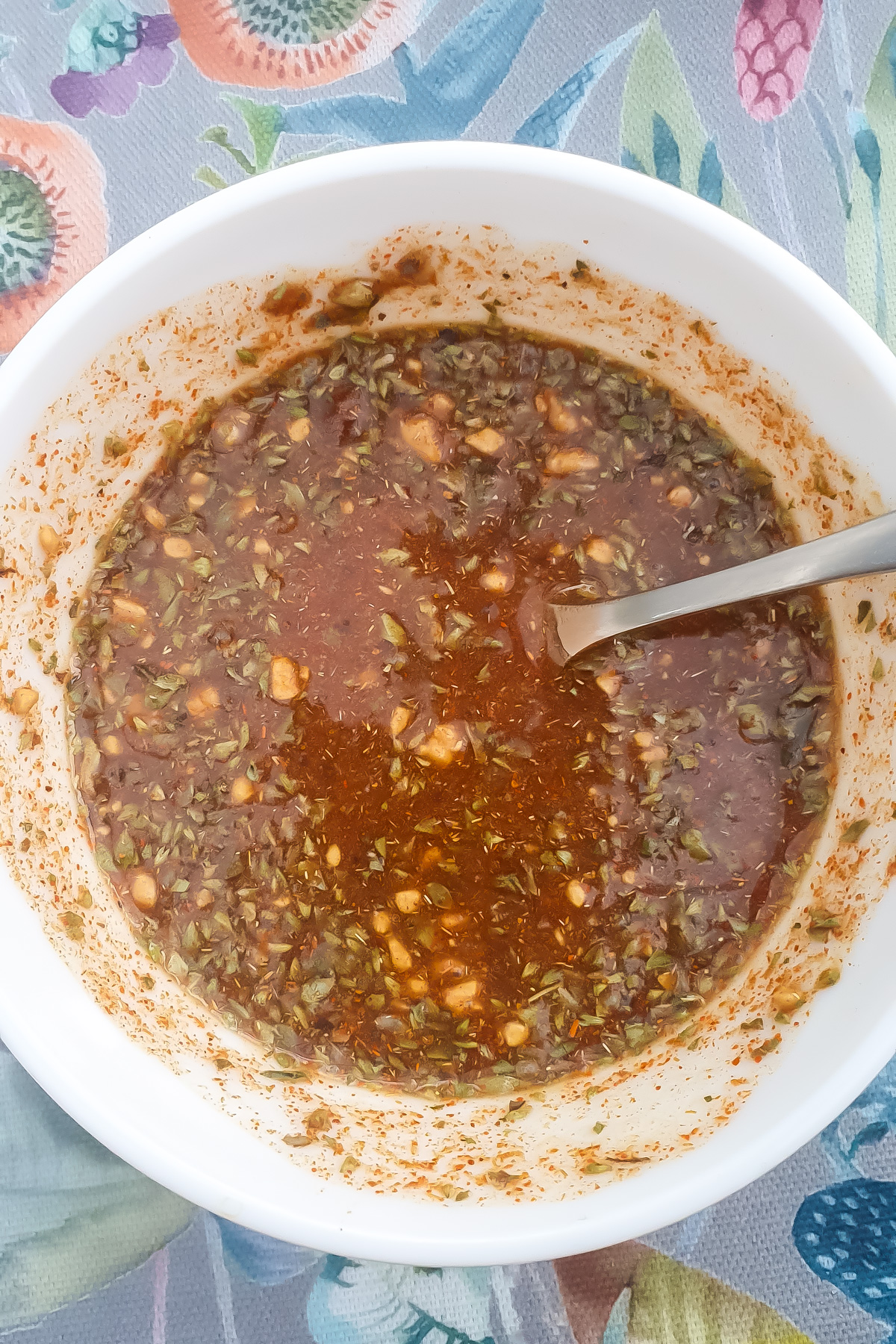 A white bowl filled with a reddish-brown sauce containing herbs, with a metal spoon inside, on a colorful patterned tablecloth.