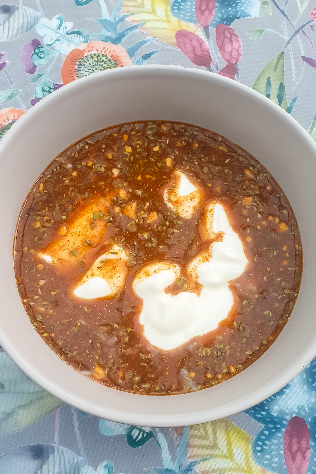 A bowl of red sauce and Greek yogurt sits on a colorful floral-patterned tablecloth.