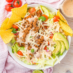 A bowl of Healthy Taco Salad with ground beef, shredded cheese, cherry tomatoes, avocado slices, tortilla chips, lettuce, and dressing on the side.