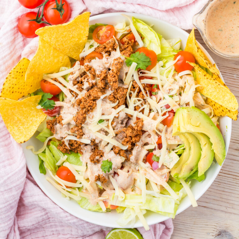 A bowl of Healthy Taco Salad with ground beef, shredded cheese, cherry tomatoes, avocado slices, tortilla chips, lettuce, and dressing on the side.