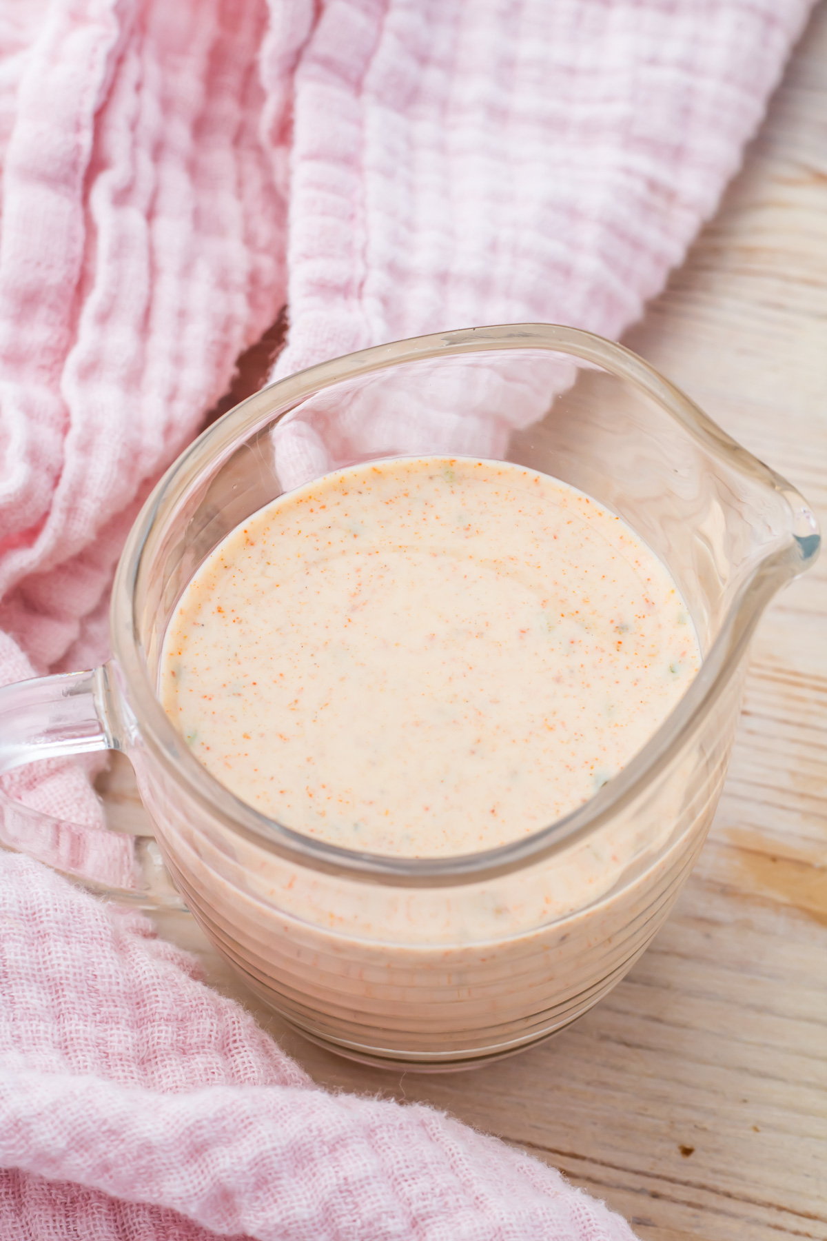 A glass measuring cup filled with a creamy, Taco Salad dressing sits on a light wooden surface next to a pink textured cloth.