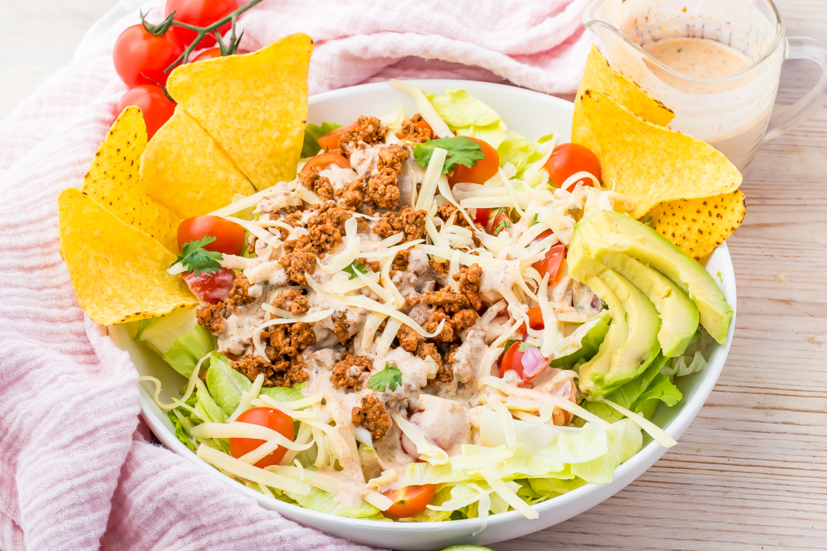A bowl of Healthy Taco Salad with ground meat, shredded cheese, avocado slices, cherry tomatoes, lettuce, tortilla chips, and a cup of creamy dressing on the side.