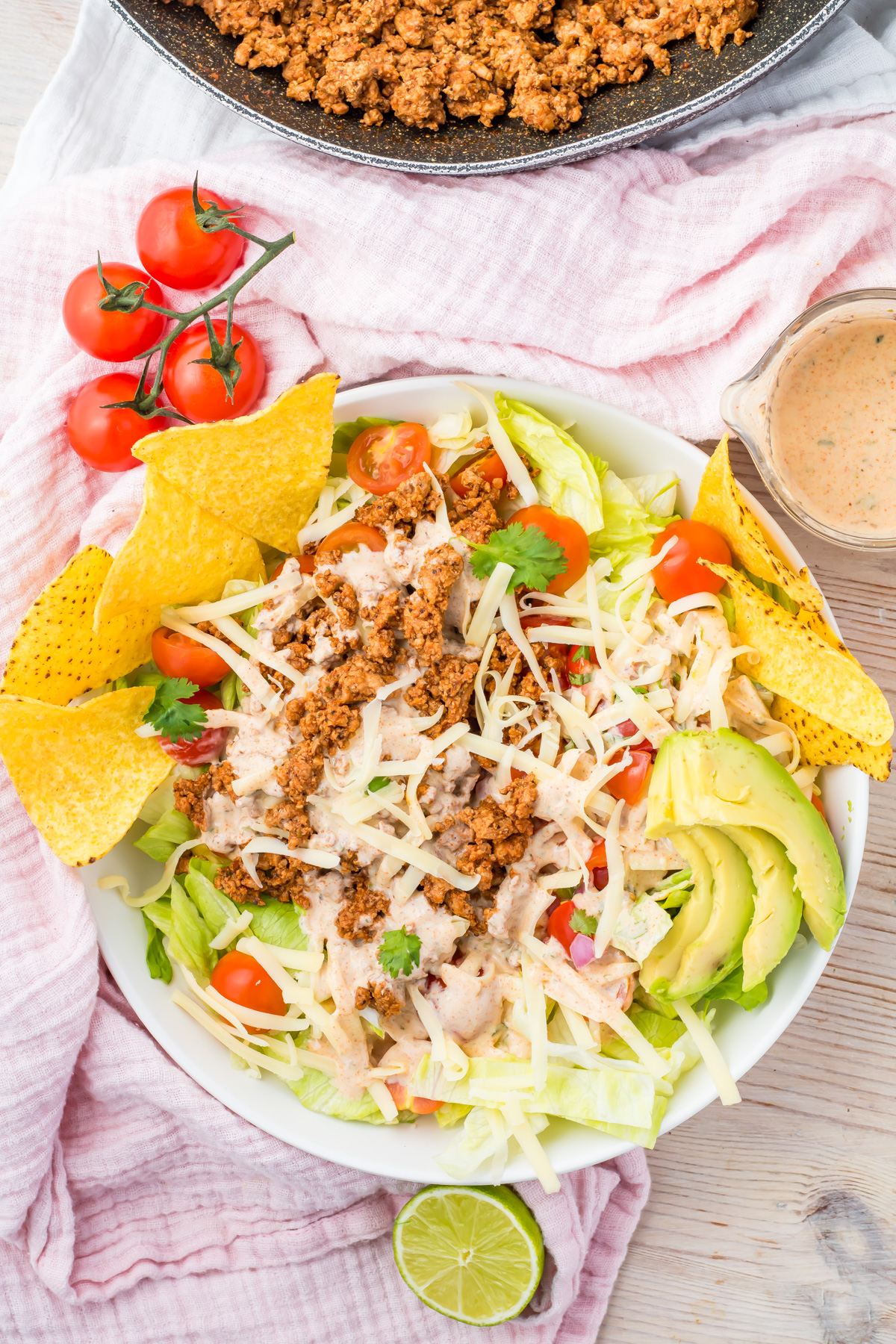 A bowl of Healthy Taco Salad with lettuce, ground meat, shredded cheese, cherry tomatoes, avocado slices, tortilla chips, and creamy dressing. A pan of meat, tomatoes, and a lime are nearby.