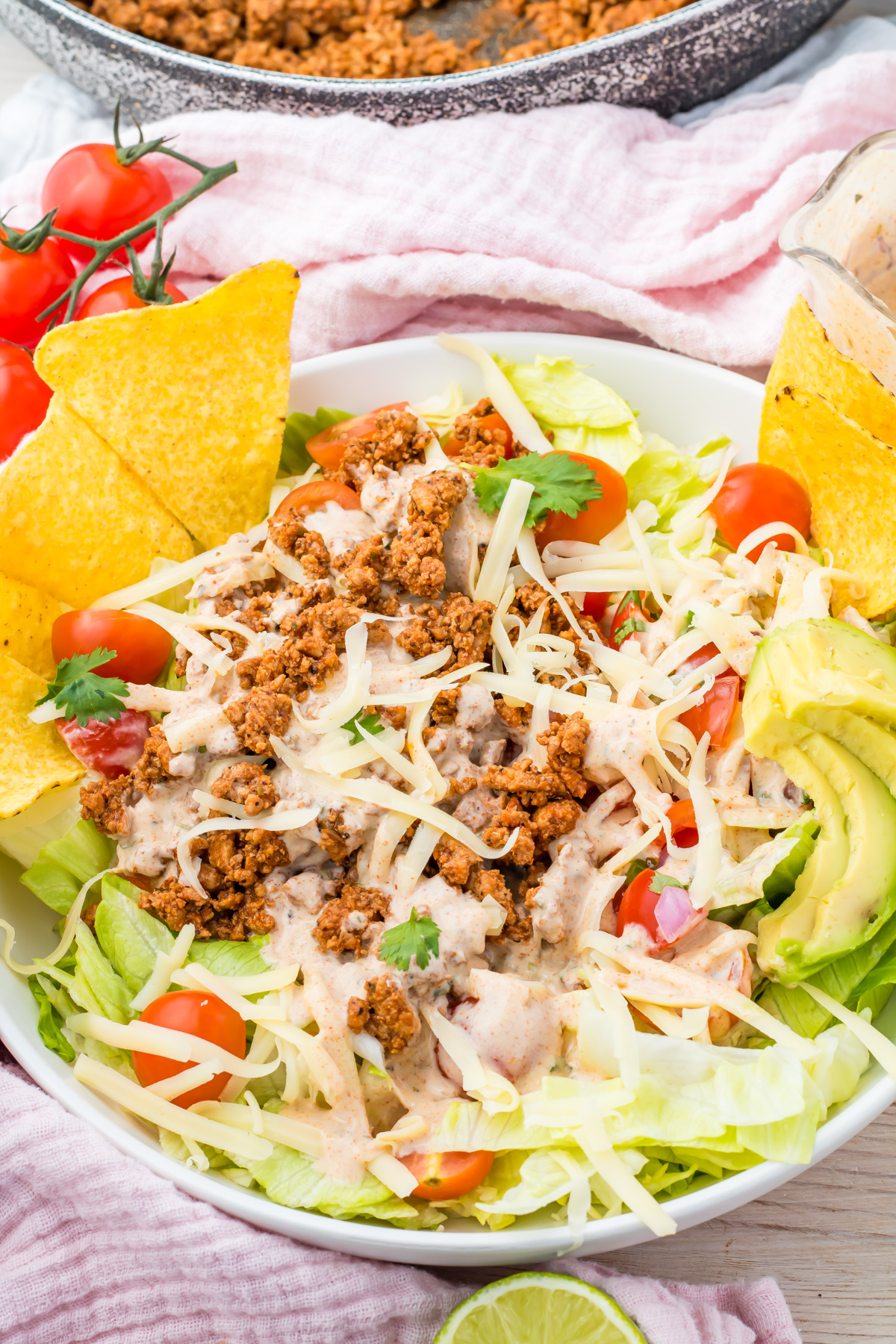 A Healthy Taco Salad with ground meat, shredded cheese, crisp lettuce, avocado slices, cherry tomatoes, tortilla chips, and creamy dressing served in a white bowl.