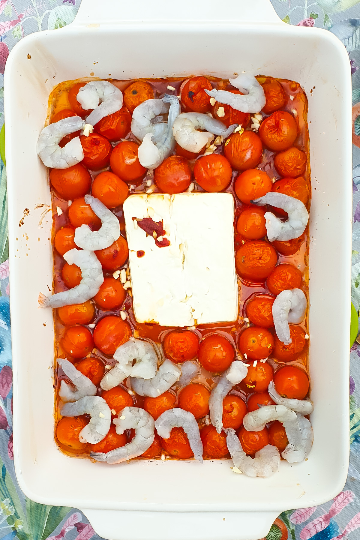 Raw shrimp arranged around cherry tomatoes and a block of feta cheese in a white baking dish.