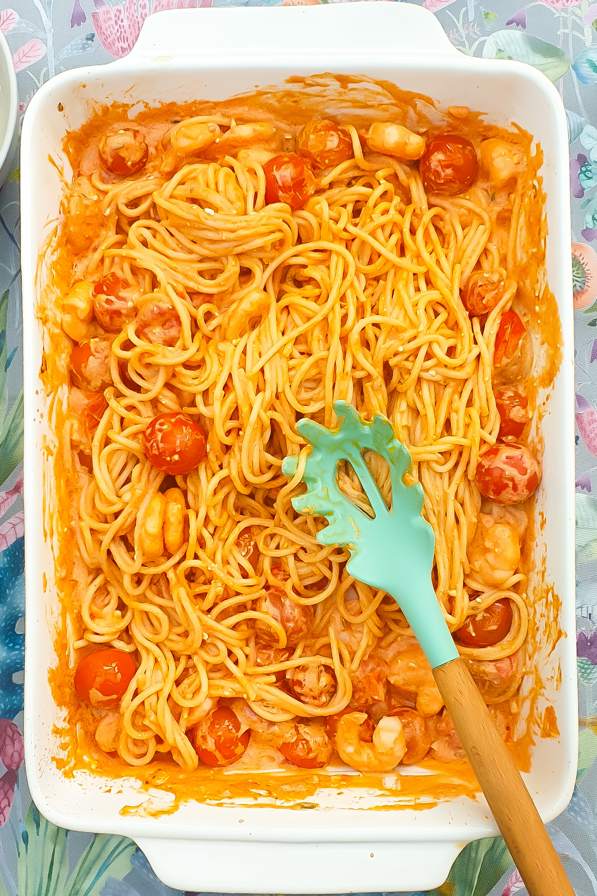 A white baking dish filled with Shrimp Pasta Bake—spaghetti, cherry tomatoes, shrimp, and creamy orange sauce. A green pasta server rests on top of the noodles.