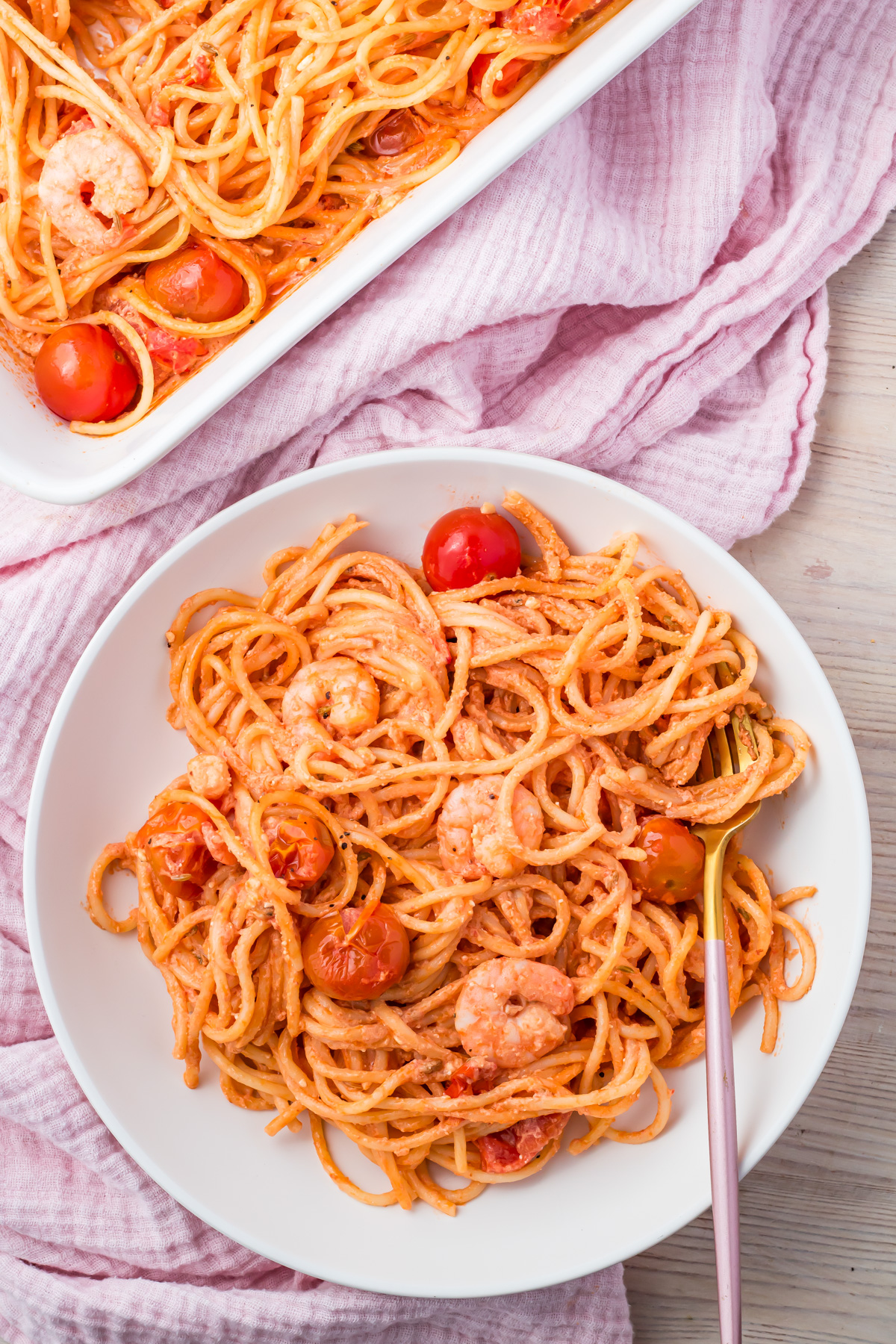 A white plate of Shrimp Pasta Bake with cherry tomatoes in a red sauce sits next to a baking dish, on a light pink cloth. A fork rests on the plate.