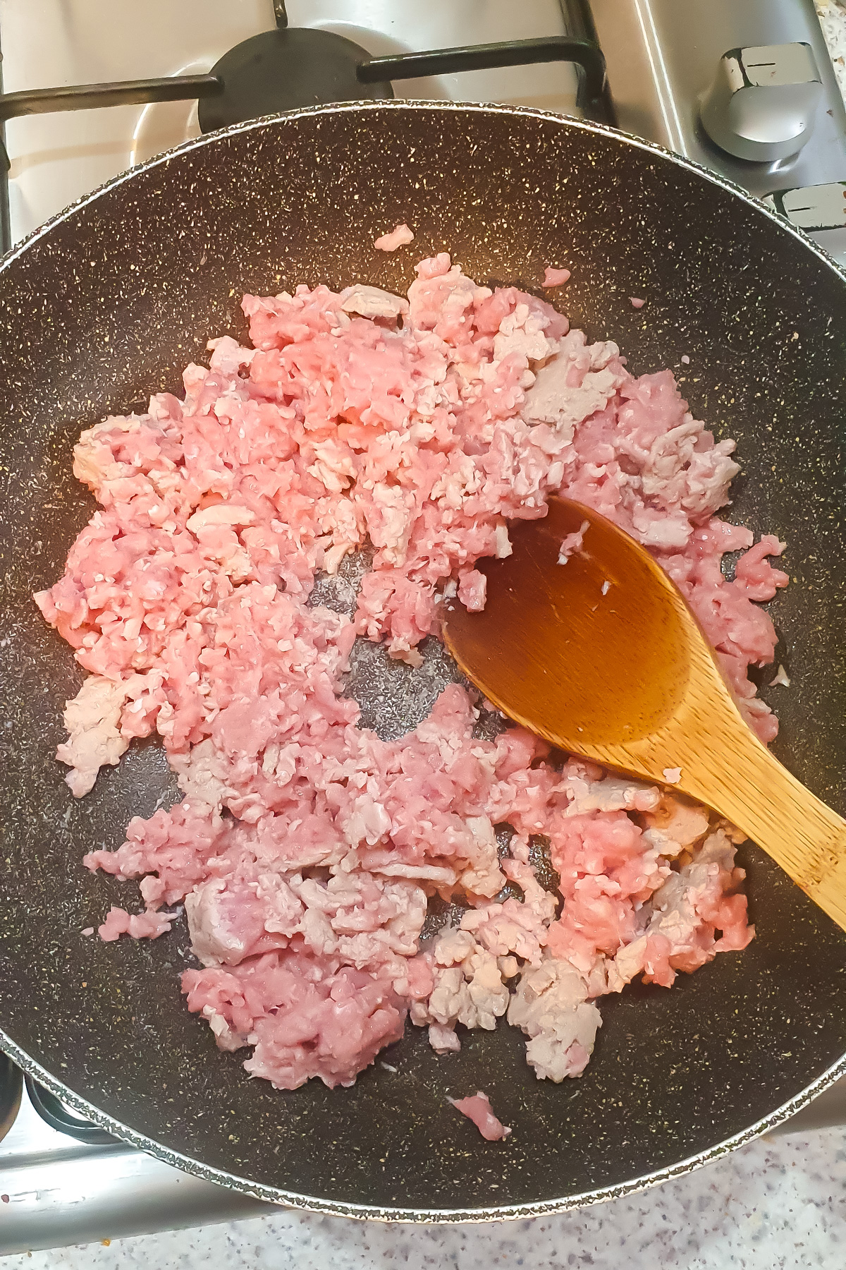 Minced meat being cooked in a frying pan on a stovetop, with a wooden spoon stirring.