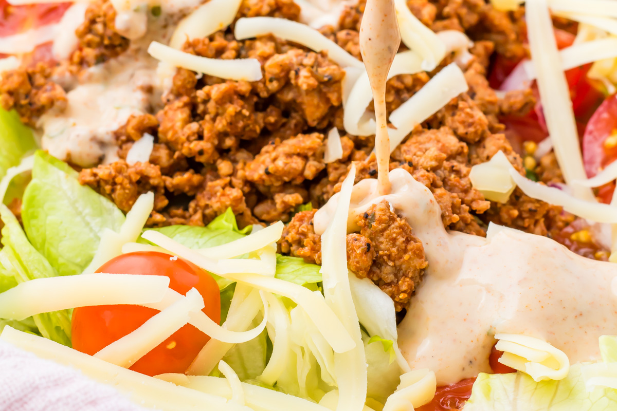 Close-up of a taco salad with ground meat, shredded cheese, lettuce, and cherry tomatoes, finished with a generous drizzle of creamy Taco Salad Dressing on top.
