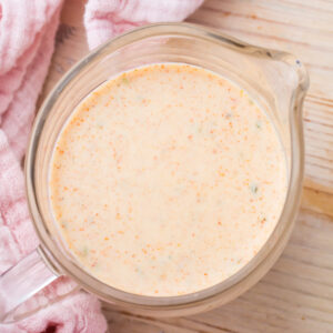 A glass measuring cup filled with a creamy, orange-colored taco salad dressing sits on a light wooden surface next to a pink cloth.