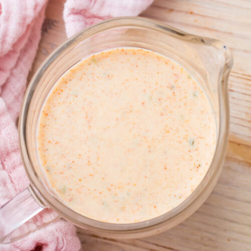 A glass measuring cup filled with a creamy, orange-colored taco salad dressing sits on a light wooden surface next to a pink cloth.