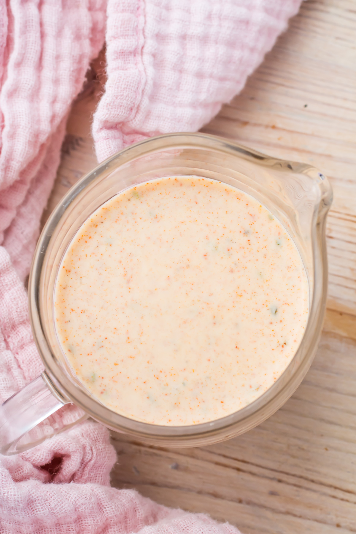 A glass measuring cup filled with creamy, orange-colored Taco Salad Dressing sits on a wooden surface next to a pink textured cloth.