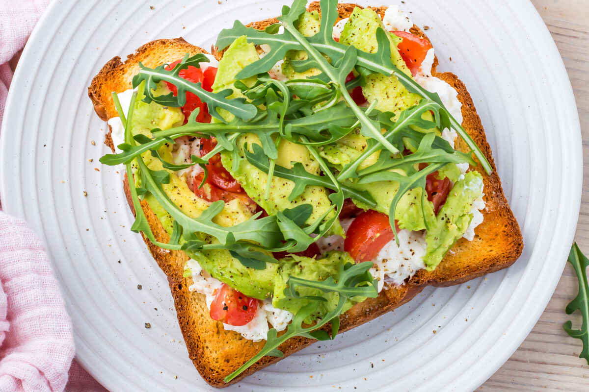 A slice of toast topped with diced avocado, cherry tomatoes, arugula, and cottage cheese rests on a white plate. Beside it, a half avocado and cherry tomatoes are arranged on a pink cloth.