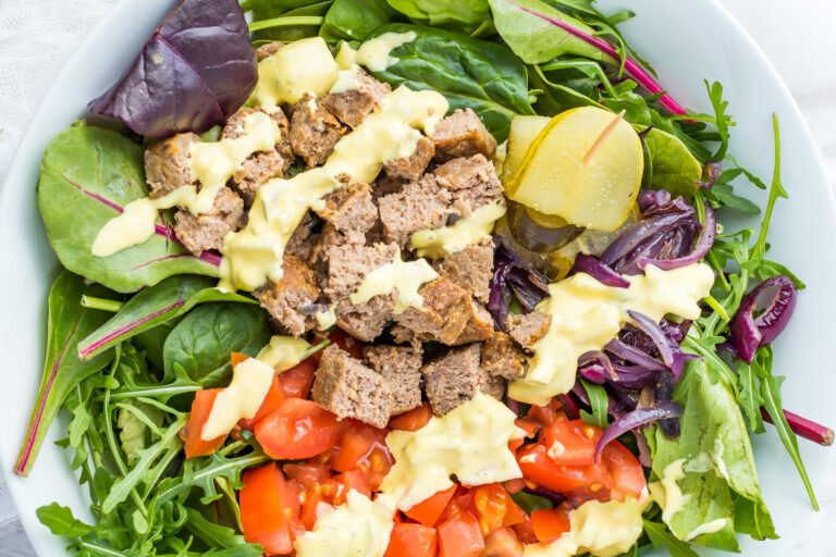 A salad bowl with mixed greens, diced tomatoes, pickles, red onions, cubed cooked meat, and a drizzle of yellow dressing—perfect for those seeking delicious Protein Bowl Recipes.