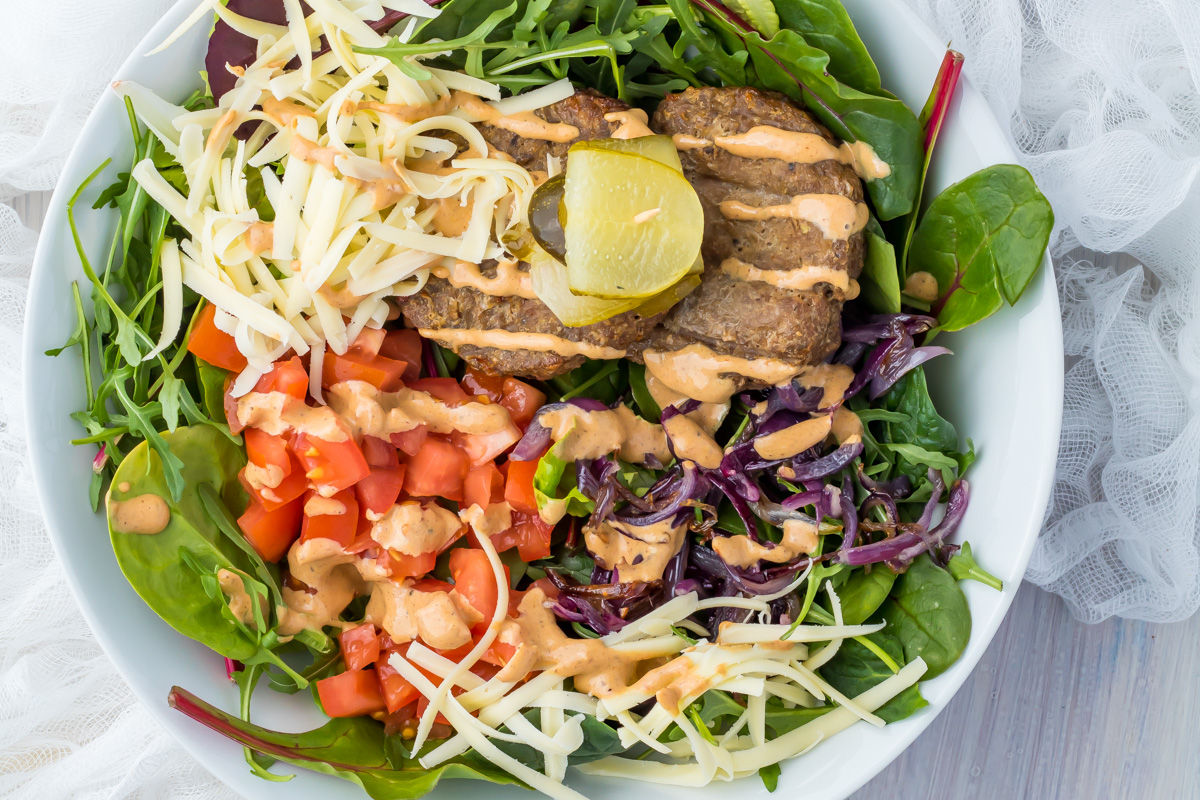 A cheeseburger protein bowl recipe featuring a vibrant mix of chopped tomatoes, shredded cheese, greens, purple cabbage, two grilled patties, a lime wedge, a green olive, and a drizzle of creamy dressing.