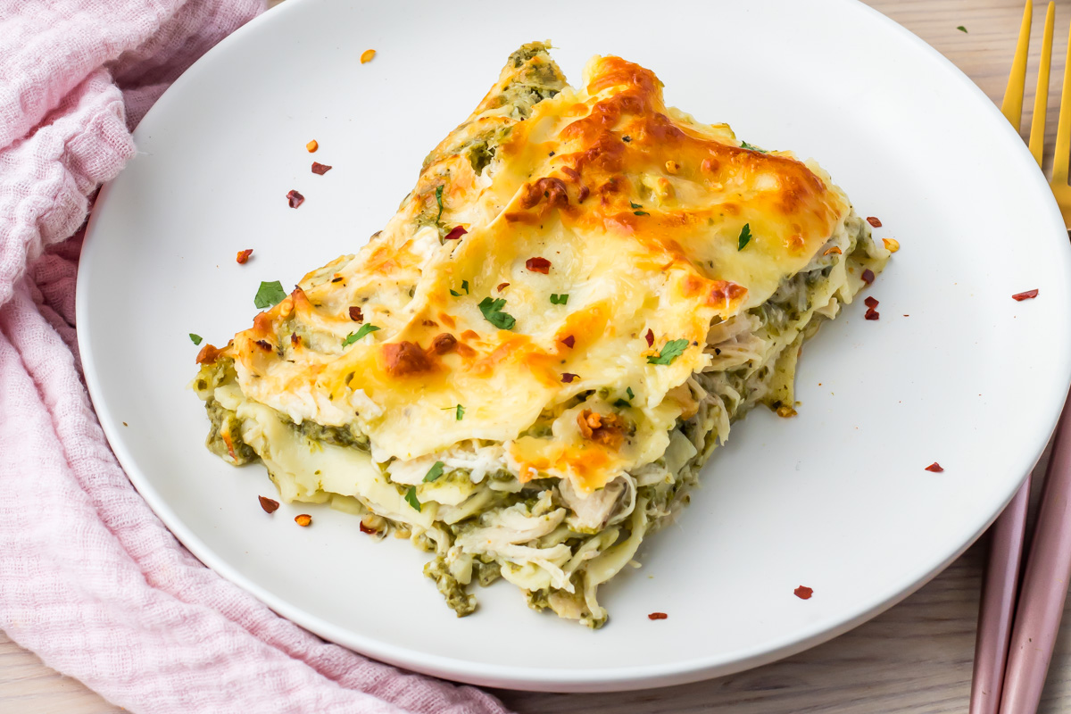 A slice of baked chicken spinach lasagna with melted cheese and herbs is served on a white plate next to a pink cloth napkin and gold-toned fork.