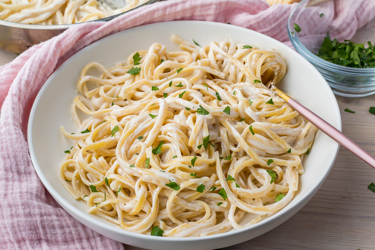 A bowl of fettuccine pasta coated in a creamy white sauce made with cottage cheese, garnished with chopped parsley, sits on a table with a fork and a pink cloth nearby.