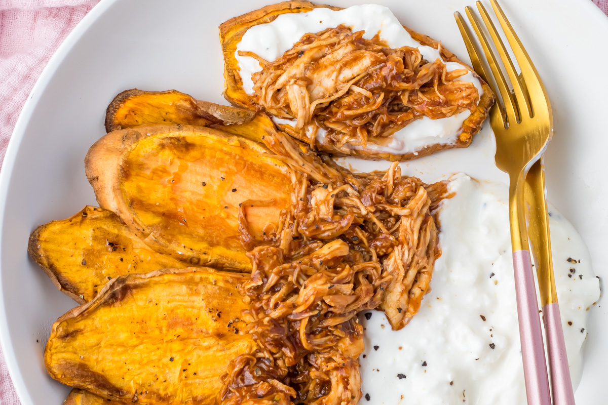 Sliced roasted sweet potatoes topped with shredded barbecue chicken and a dollop of cottage cheese, served on a white plate with a gold and pink fork.