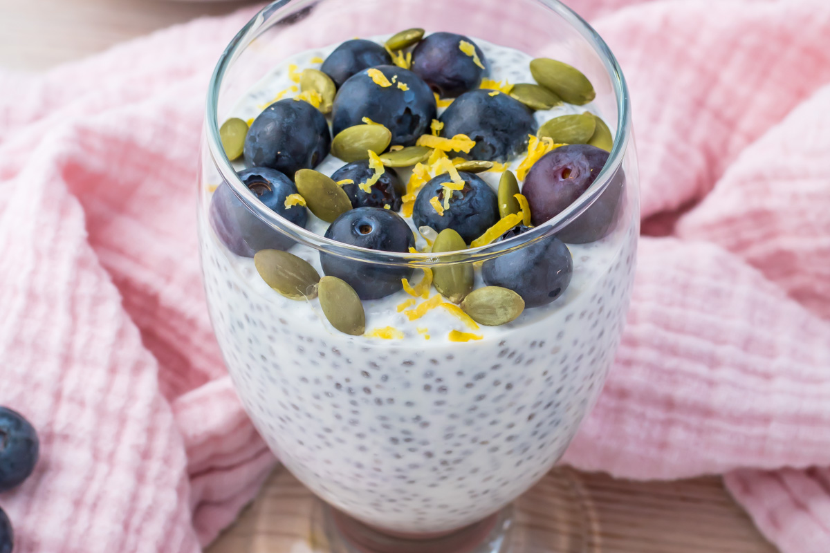 A glass of cottage cheese chia pudding topped with fresh blueberries, pumpkin seeds, and lemon zest sits on a pink cloth.