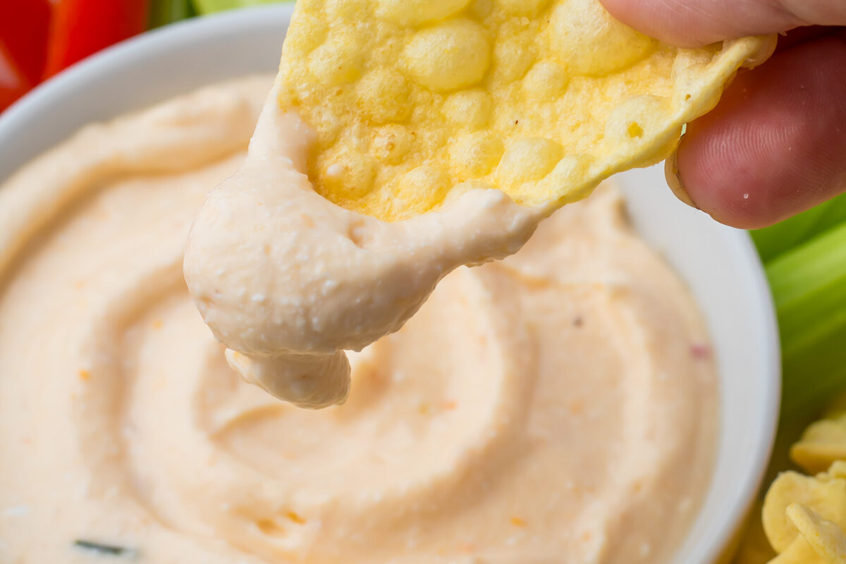 A hand dips a crispy chip into a bowl of creamy Cottage Cheese Queso, surrounded by celery sticks and red bell pepper slices.