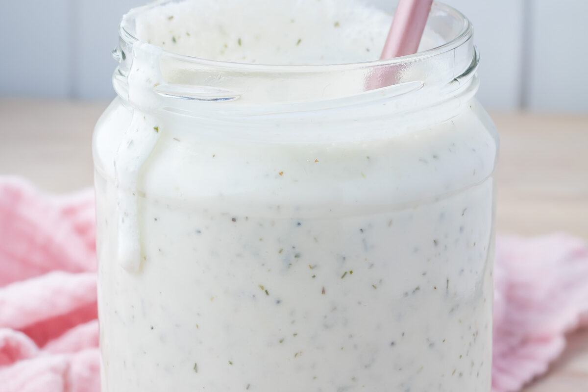 A glass jar filled with creamy, speckled cottage cheese ranch dressing, with a pink spoon inside. The jar rests on a wooden surface, accompanied by a pink cloth.