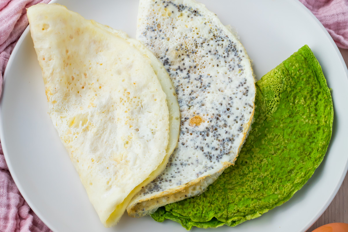 Three egg white wraps are arranged on a white plate: one plain, one with chia seeds, and one green, made with spinach.