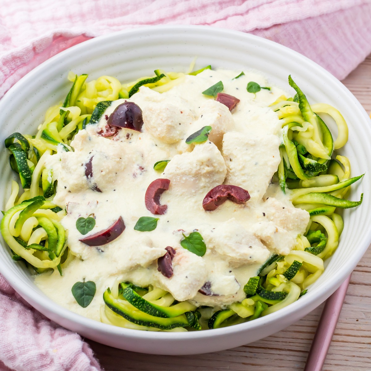 A white bowl of zucchini noodles topped with Greek Yogurt Chicken, creamy white sauce, sliced black olives, and fresh herbs sits next to a pink cloth.