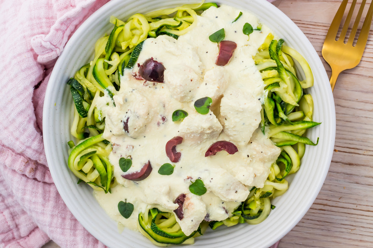 A bowl of zucchini noodles topped with creamy Greek Yogurt Chicken, sliced olives, fresh herbs, and a rich white sauce, with a gold fork and pink cloth nearby.