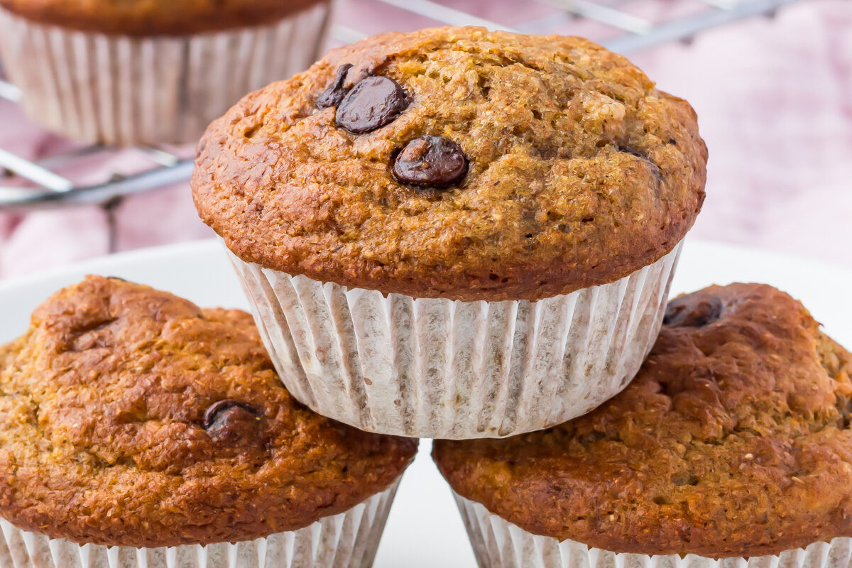 Three healthy banana muffins, studded with chocolate chips, stacked on a white plate, with more muffins cooling on a rack in the background.