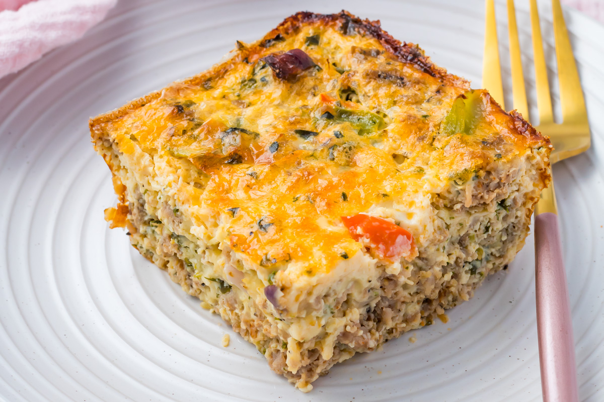 A slice of egg bake casserole featuring ground meat, eggs, vegetables, and melted cheese is served on a white plate with a gold fork beside it.