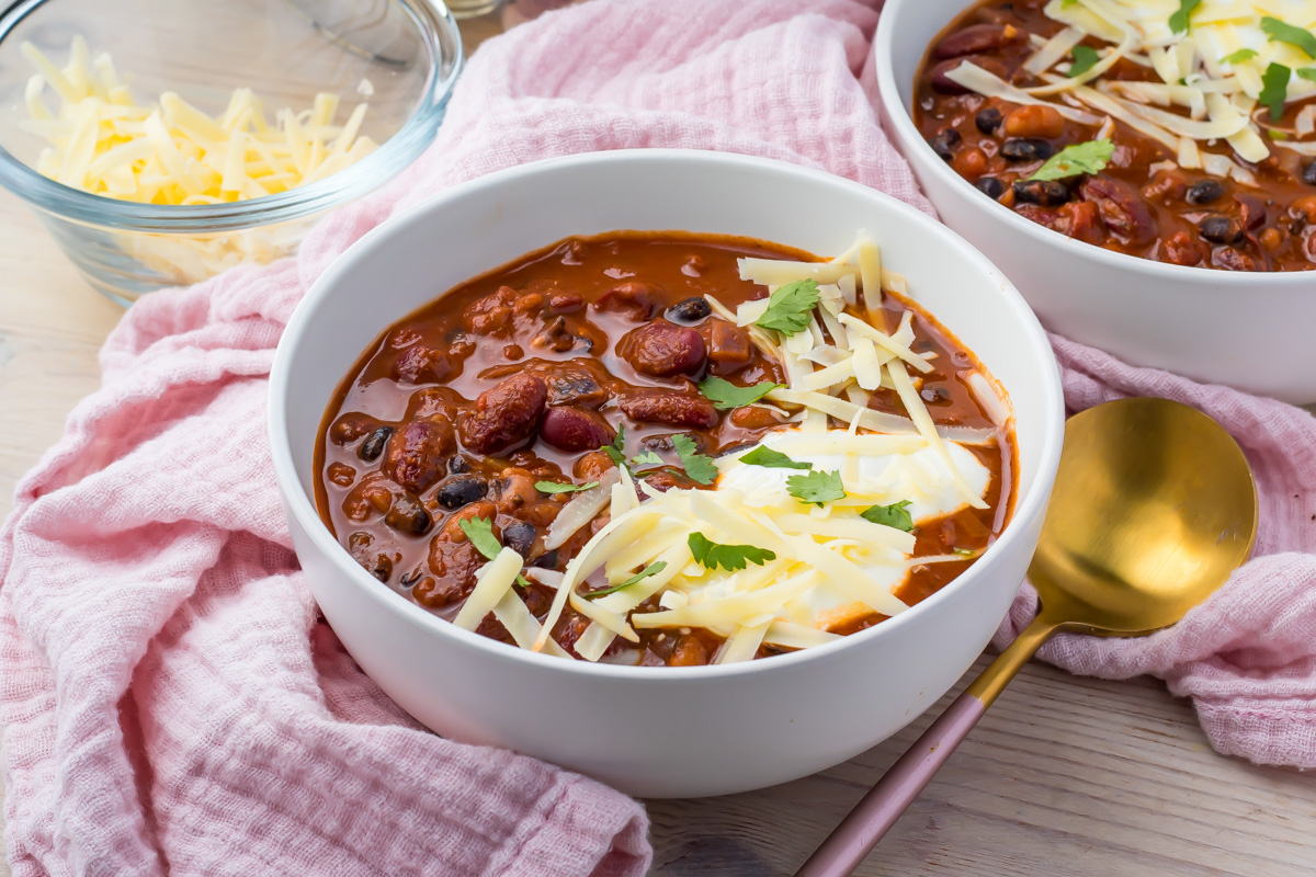 A white bowl of high protein vegan chili topped with plant based shredded cheese and cilantro sits on a pink cloth, with a gold spoon and a bowl of cheese in the background.