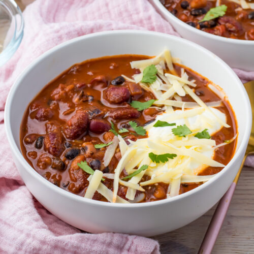 A white bowl filled with high protein vegan chili topped with dairy free shredded cheese, sour cream, and cilantro, placed on a pink cloth napkin.