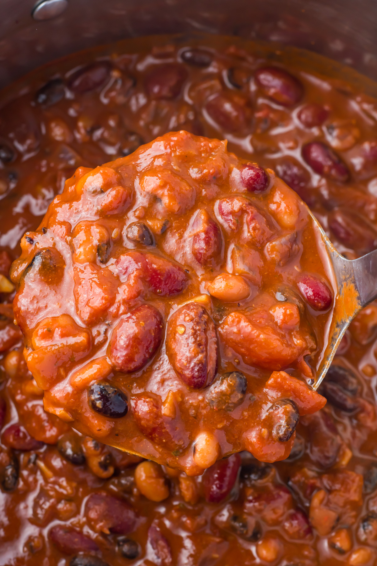 A close-up of a ladle filled with thick, chunky high protein vegan chili containing beans and tomatoes over a pot of chili.