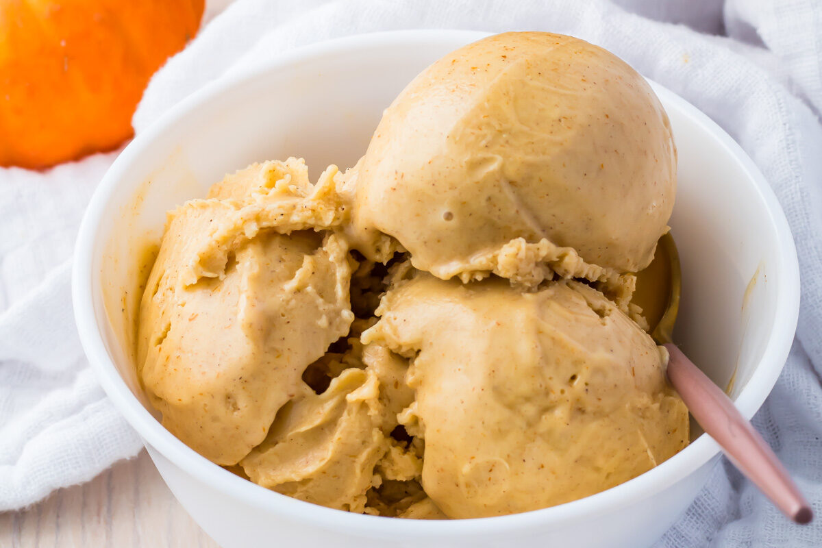 A bowl of three scoops of protein pumpkin ice cream rests on a white cloth, with a small pumpkin and an ice cream container in the background.