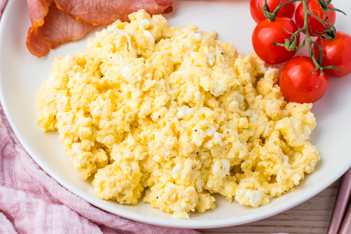 A plate with scrambled eggs enriched with creamy cottage cheese, cherry tomatoes on the vine, and crispy bacon rests on a white dish, placed on a pink cloth. The perfect breakfast recipe to start your day.