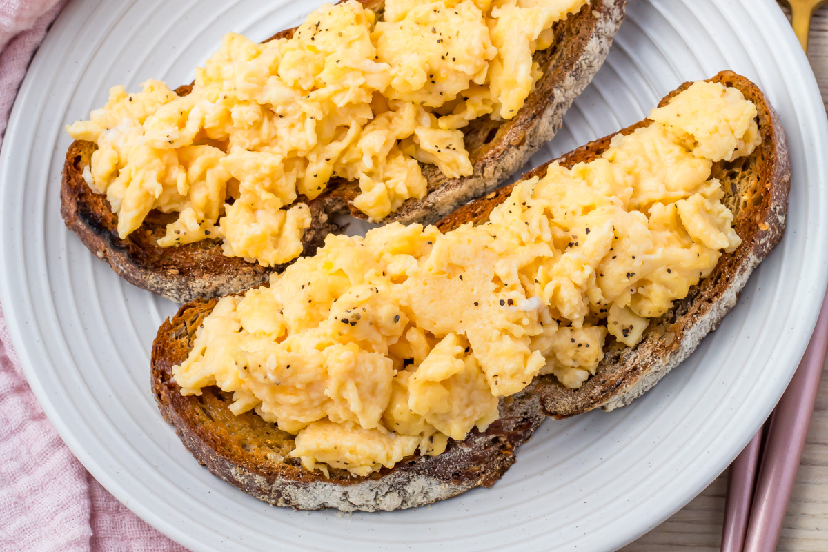 Two slices of toasted bread topped with scrambled eggs with water are served on a white plate.