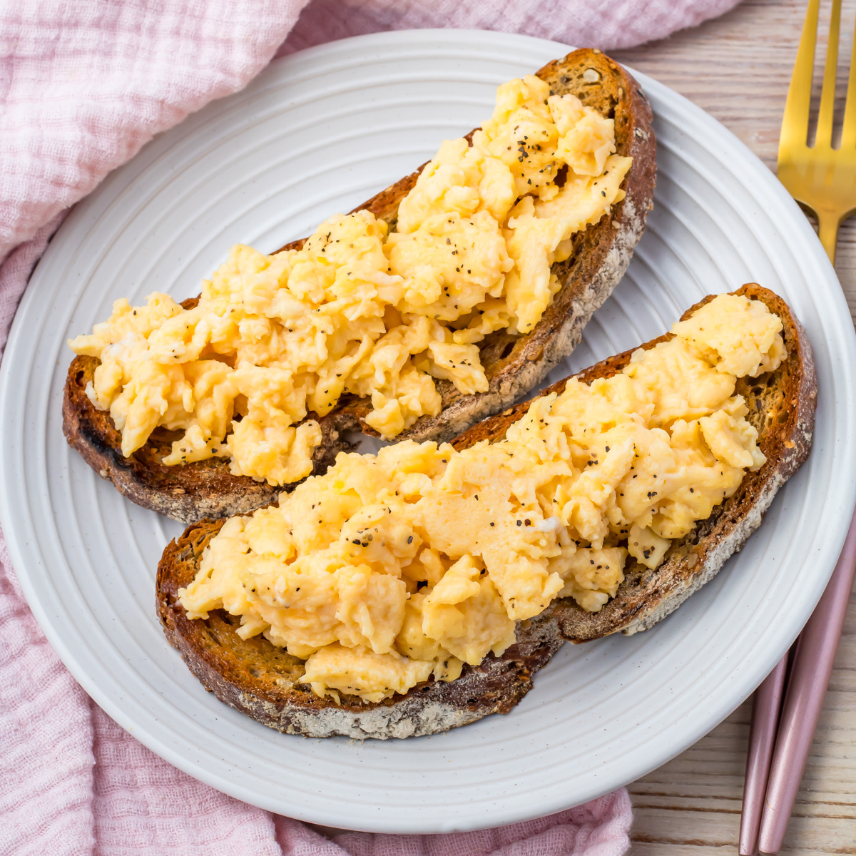 Two slices of toasted bread topped with scrambled eggs with water are served on a white plate, beside a gold fork and knife.