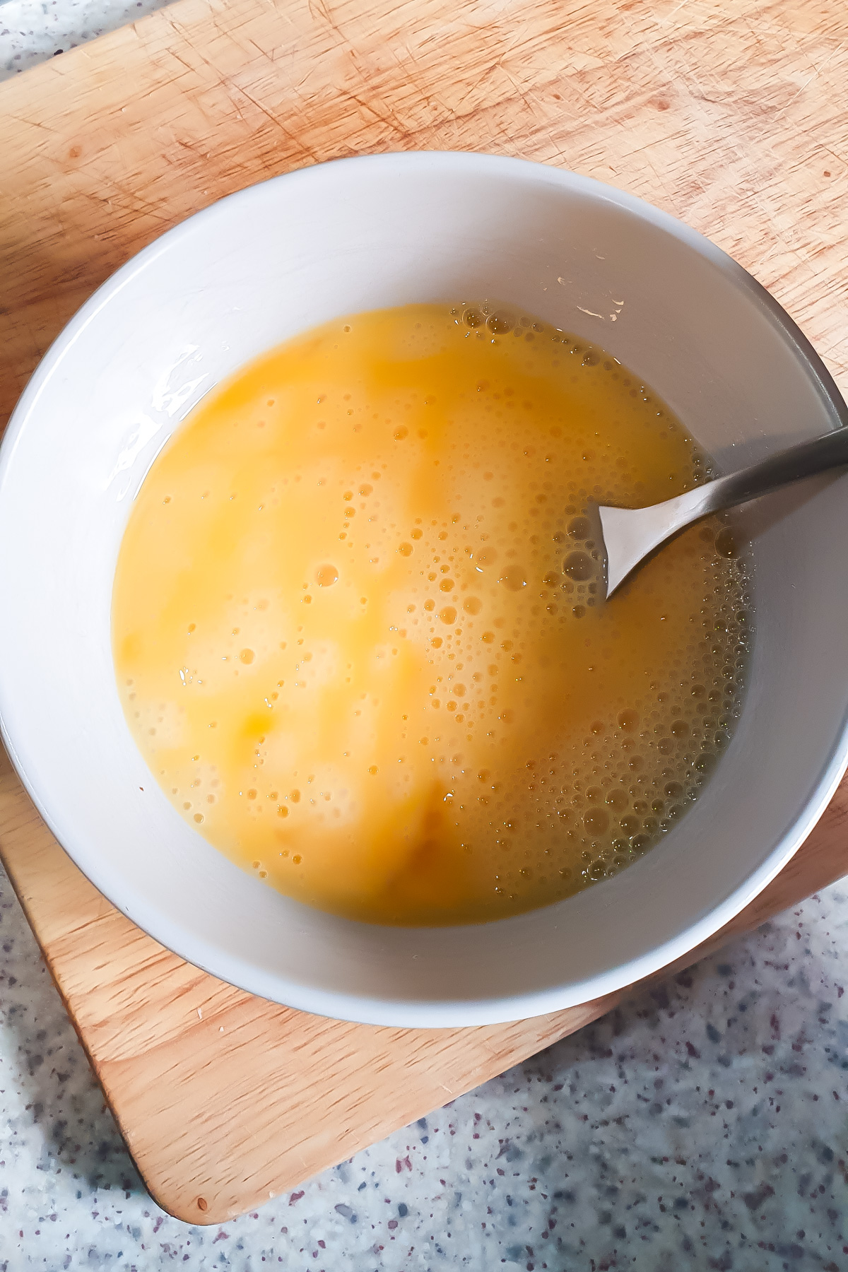A bowl of beaten eggs with a fork inside, placed on a wooden cutting board on a speckled countertop.