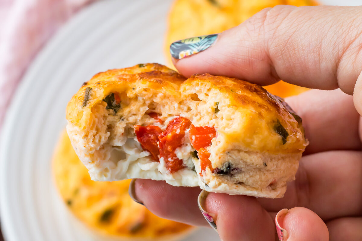 A hand holding a half-eaten starbucks egg white bite with visible pieces of red pepper and cheese, with more muffins on a white plate in the background.