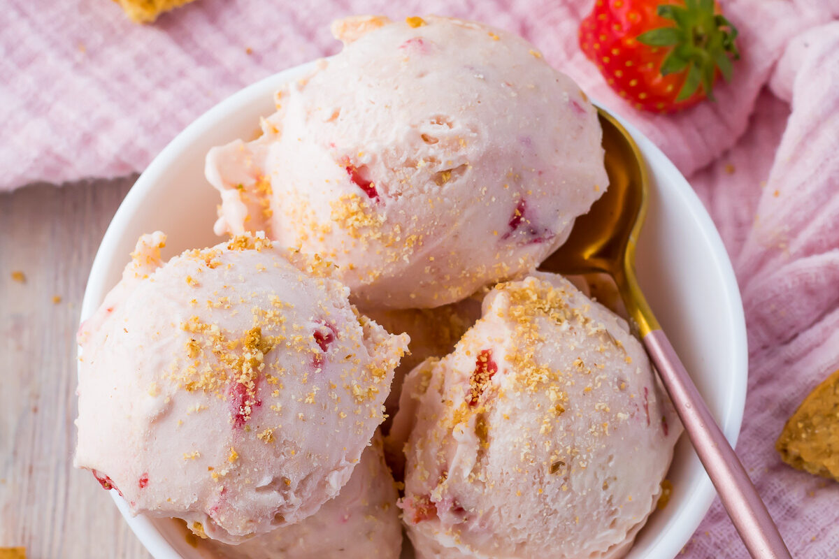 Strawberry cottage cheese ice cream in a bowl beside a pink cloth with strawberries and cookies scattered around.