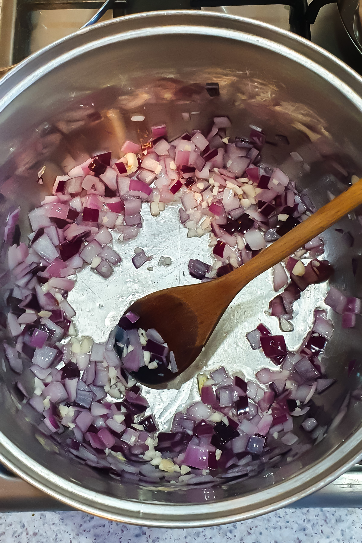 Chopped red onions and garlic being sautéed in a stainless steel pot with a wooden spoon on a stovetop—the perfect aromatic base for a high protein vegan chili.