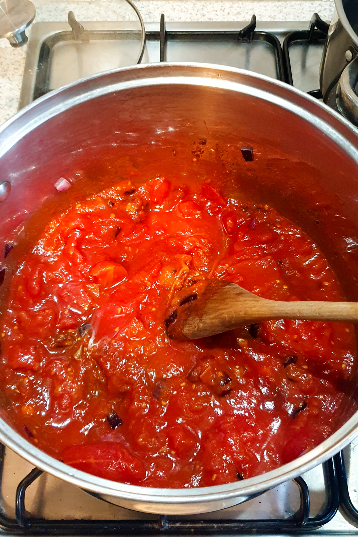 A stainless steel pot on a gas stove simmers a vegan chili tomato sauce with visible chunks of tomatoes and onions; a wooden spoon rests inside the pot.